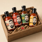 Four sauce bottles in a wooden gift box with crumpled brown paper. The bottles have black caps and colourful labels featuring a stylized bearded man's face, with text showing the flavours.